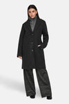Manteau long noir de JDY, porté avec un pull gris et un pantalon à carreaux.