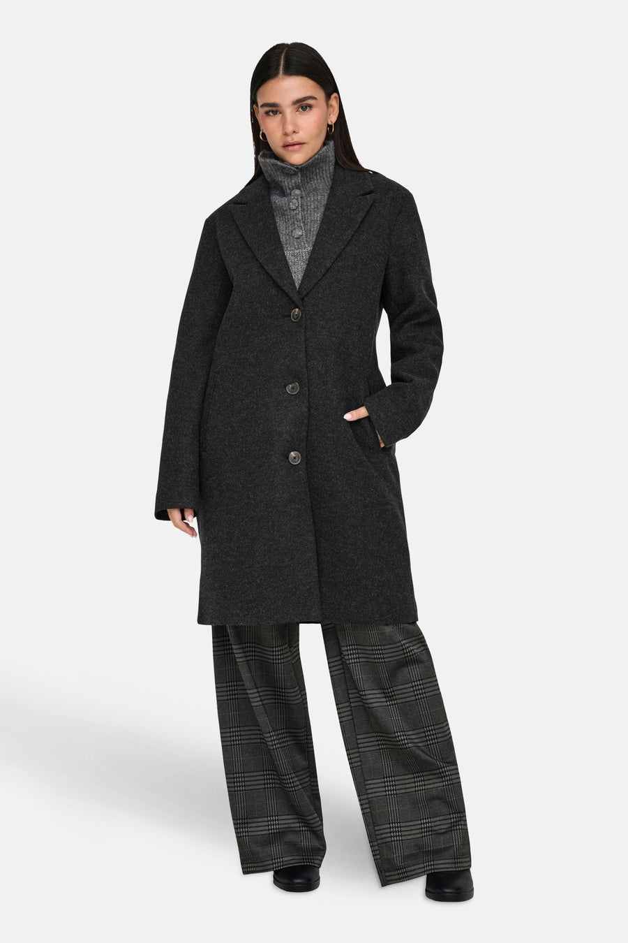 Manteau long noir de JDY, porté avec un pull gris et un pantalon à carreaux.