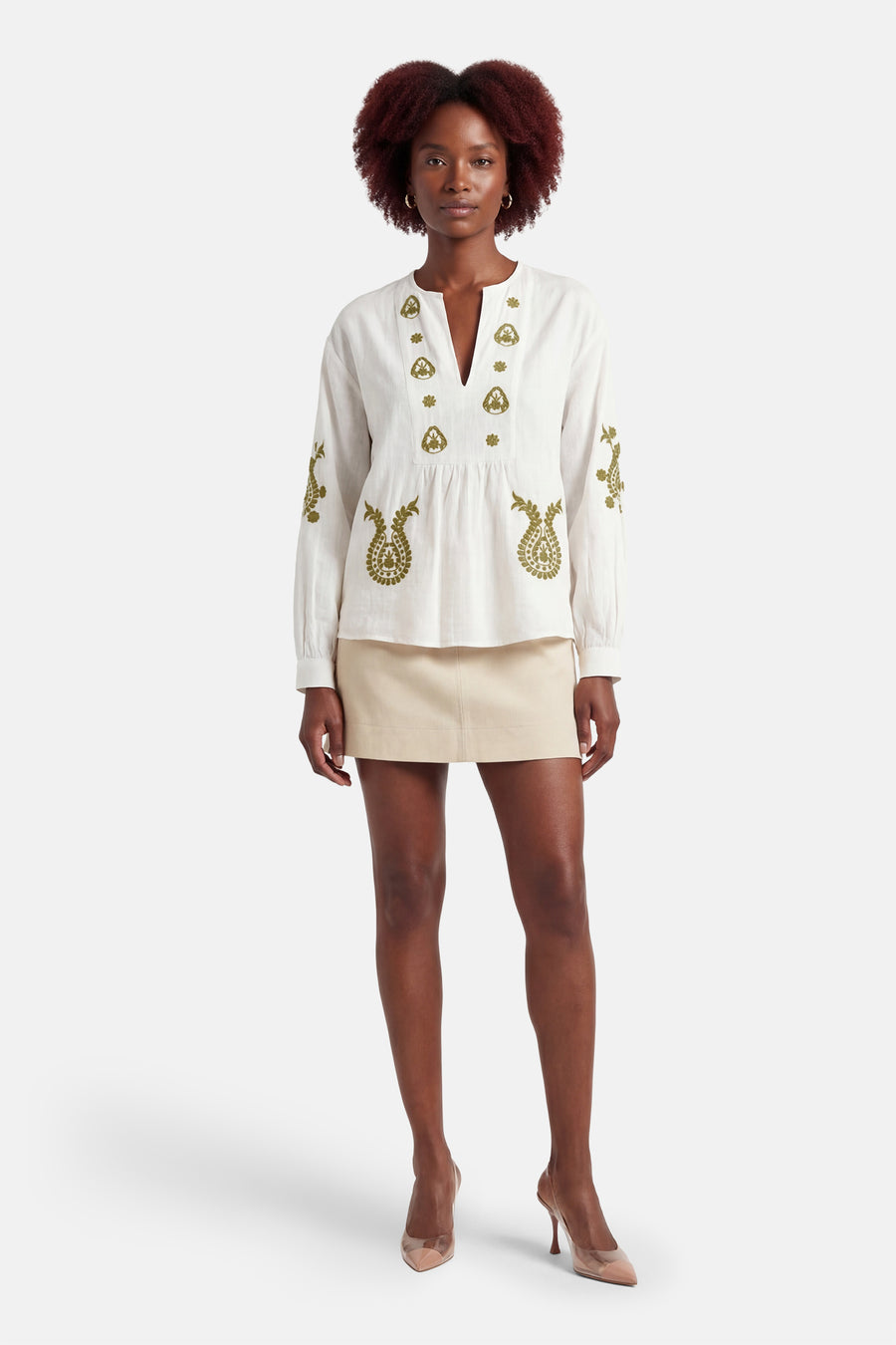 Blouse à manches longues blanche de ONLY, avec broderie florale, portée avec une minijupe beige et des escarpins à talons aiguilles.
