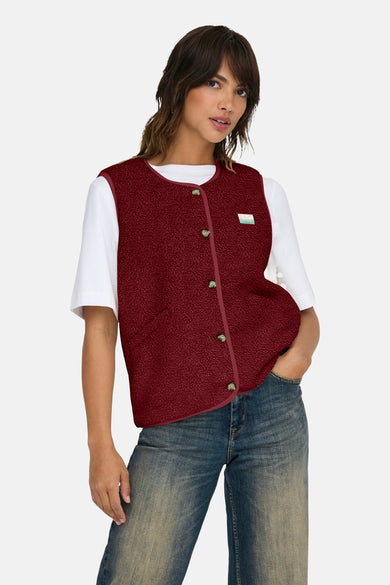 Bordeaux mouwloos vest van bouclé wol van ONLY, gedragen over een wit T-shirt en een blauwe jeans.