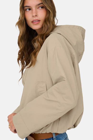 Korte beige jas van ONLY®, met capuchon en opgestikte zak, gedragen met een blauwe jeans en een bruine riem.