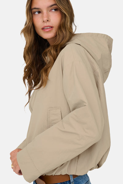 Korte beige jas van ONLY®, met capuchon en opgestikte zak, gedragen met een blauwe jeans en een bruine riem.