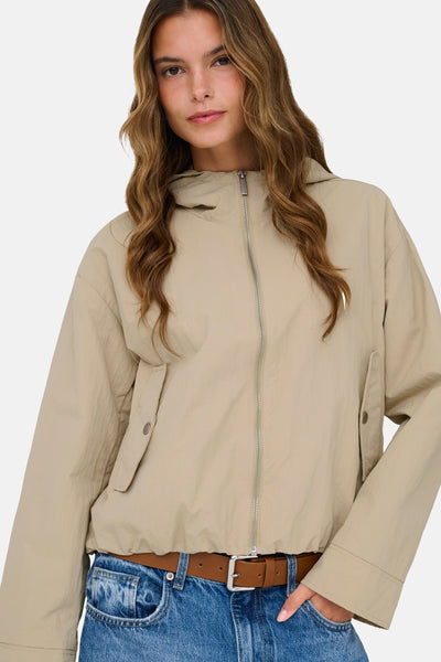 Korte beige jas van ONLY, met zilveren ritssluiting, klepzakken en een bruine riem over een blauwe jeans.