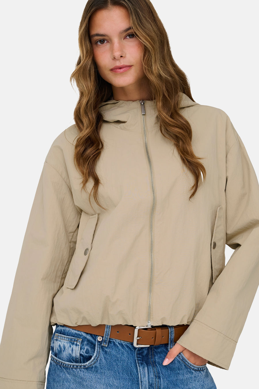 Korte beige jas van ONLY, met zilveren ritssluiting, klepzakken en een bruine riem over een blauwe jeans.