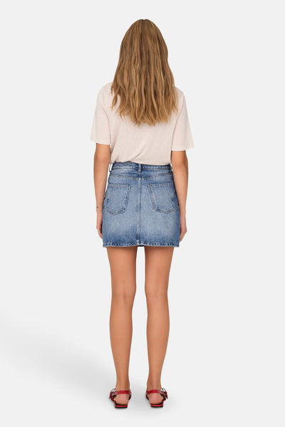Jupe courte en denim bleu clair de ONLY, avec poches arrière et ourlet effiloché, portée avec un t-shirt beige et des sandales rouges.
