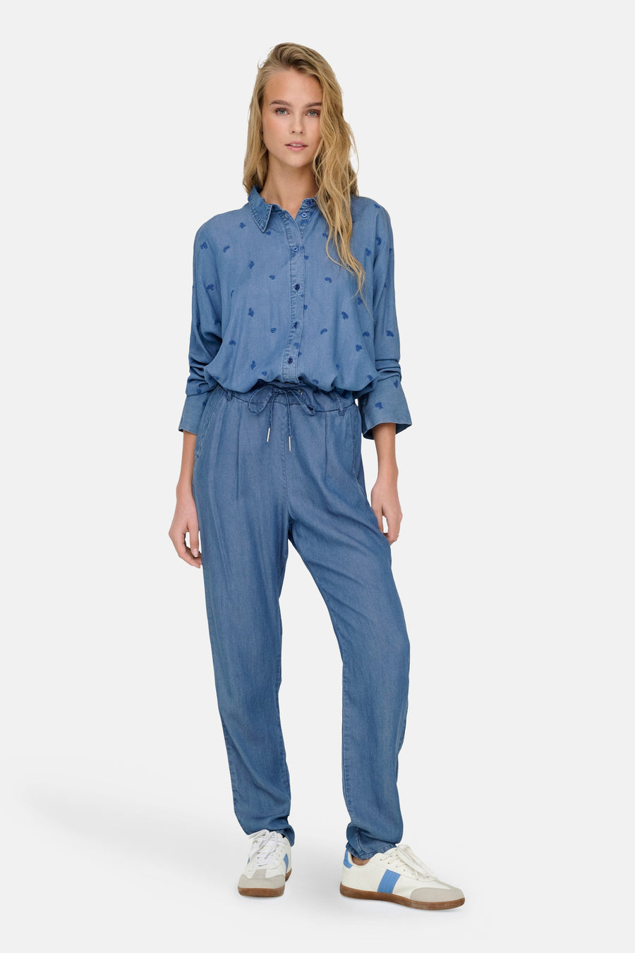 Pantalon coloré bleu denim de ONLY®, avec taille élastiquée et motif subtil, porté avec des chaussures de sport blanches.
