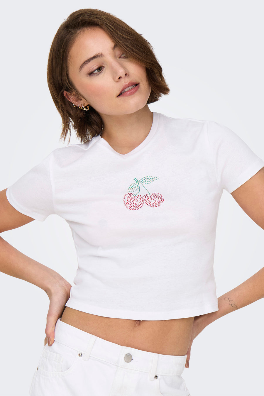 T-shirt blanc à manches courtes de ONLY, orné d'un motif de cerises en strass, porté avec un pantalon blanc.
