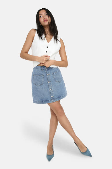 Jupe courte en denim bleu clair de ONLY®, boutonnée sur le devant avec poches plaquées, portée avec un haut blanc.