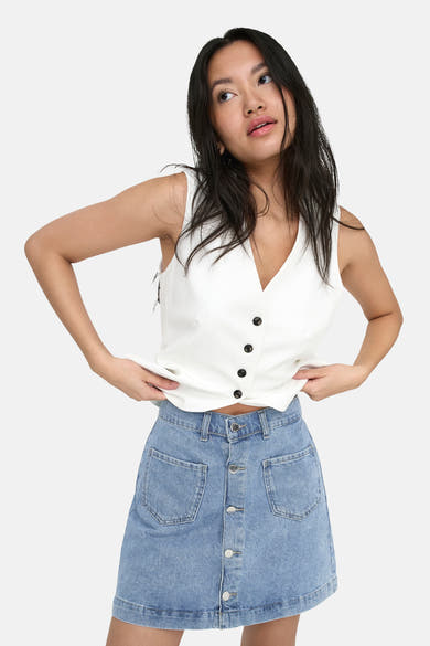 Jupe courte en denim bleu clair de ONLY® avec fermeture boutonnée et poches plaquées, portée avec un gilet blanc.