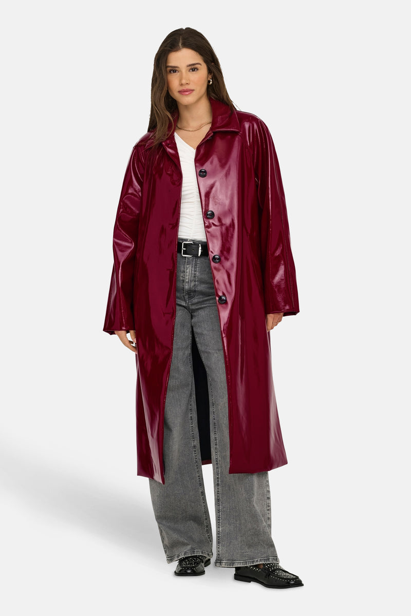 Lange bordeaux vinylen jas van ONLY, gedragen met een wit T-shirt, een grijze jeans en zwarte loafers.