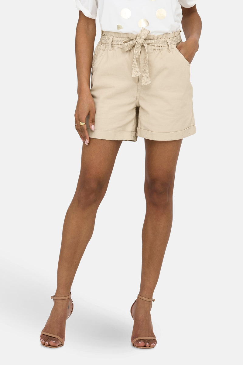 Korte beige short van JDY met een strikceintuur, gedragen met een witte top met gouden stippen en hoge hakken.
