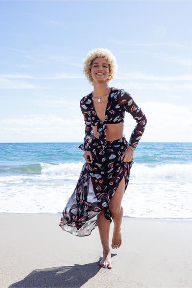 Blouse noire à manches longues de ONLY, avec un motif coquillage, portée avec une jupe assortie sur une plage.
