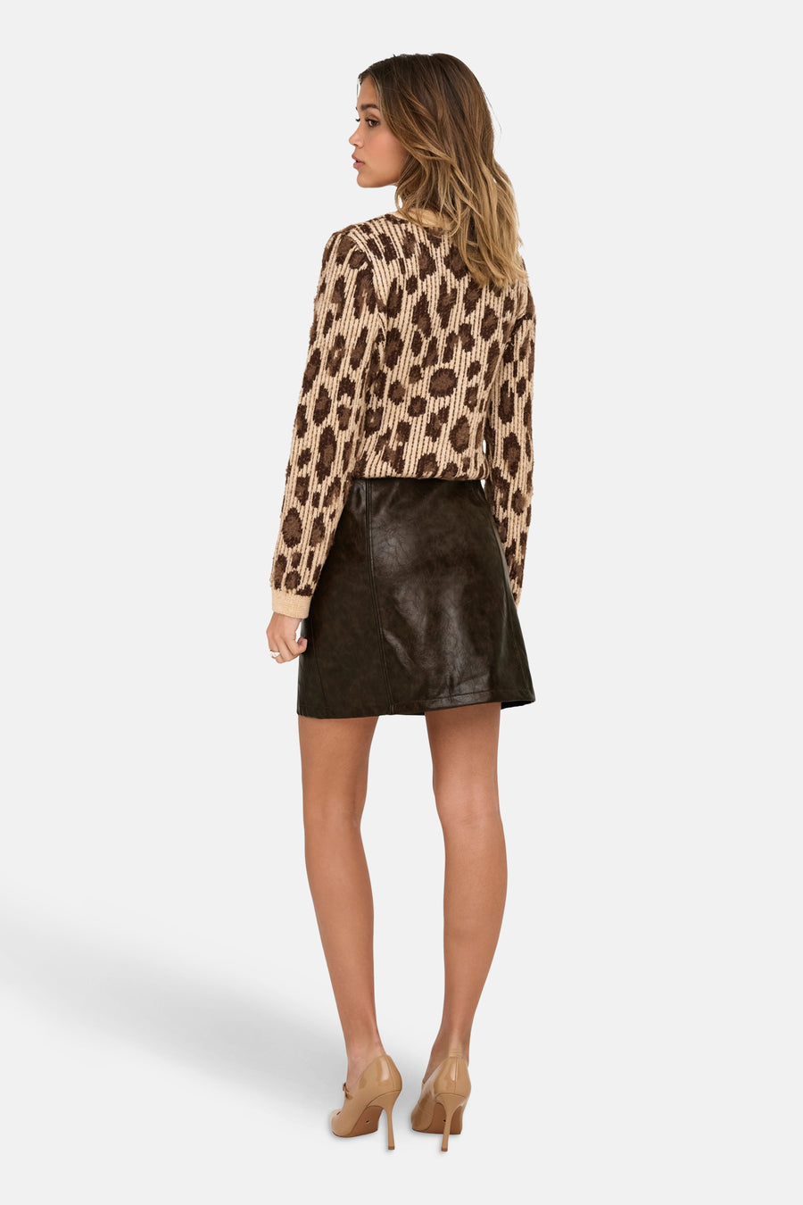 Jupe courte marron en similicuir de ONLY®, portée avec un pull à motif léopard et des talons hauts nude.
