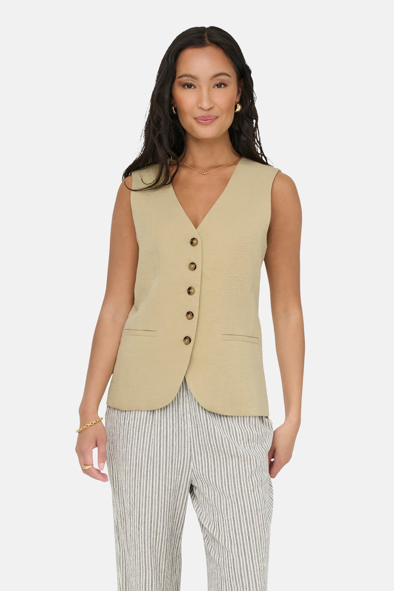Blazer écru de JDY, sans manches, avec boutons et poches, porté avec un pantalon rayé et des bijoux dorés.
