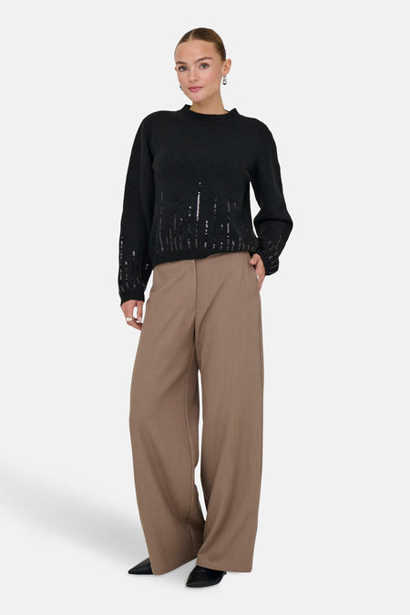 Zwarte rondehals trui van JDY, met verticale pailletten details, gedragen met een bruine broek met wijde pijpen en zwarte schoenen.
