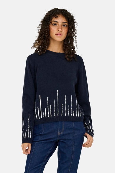 Pull à col rond bleu de JDY avec motif vertical argenté sur le devant et les manches, porté avec un jean.
