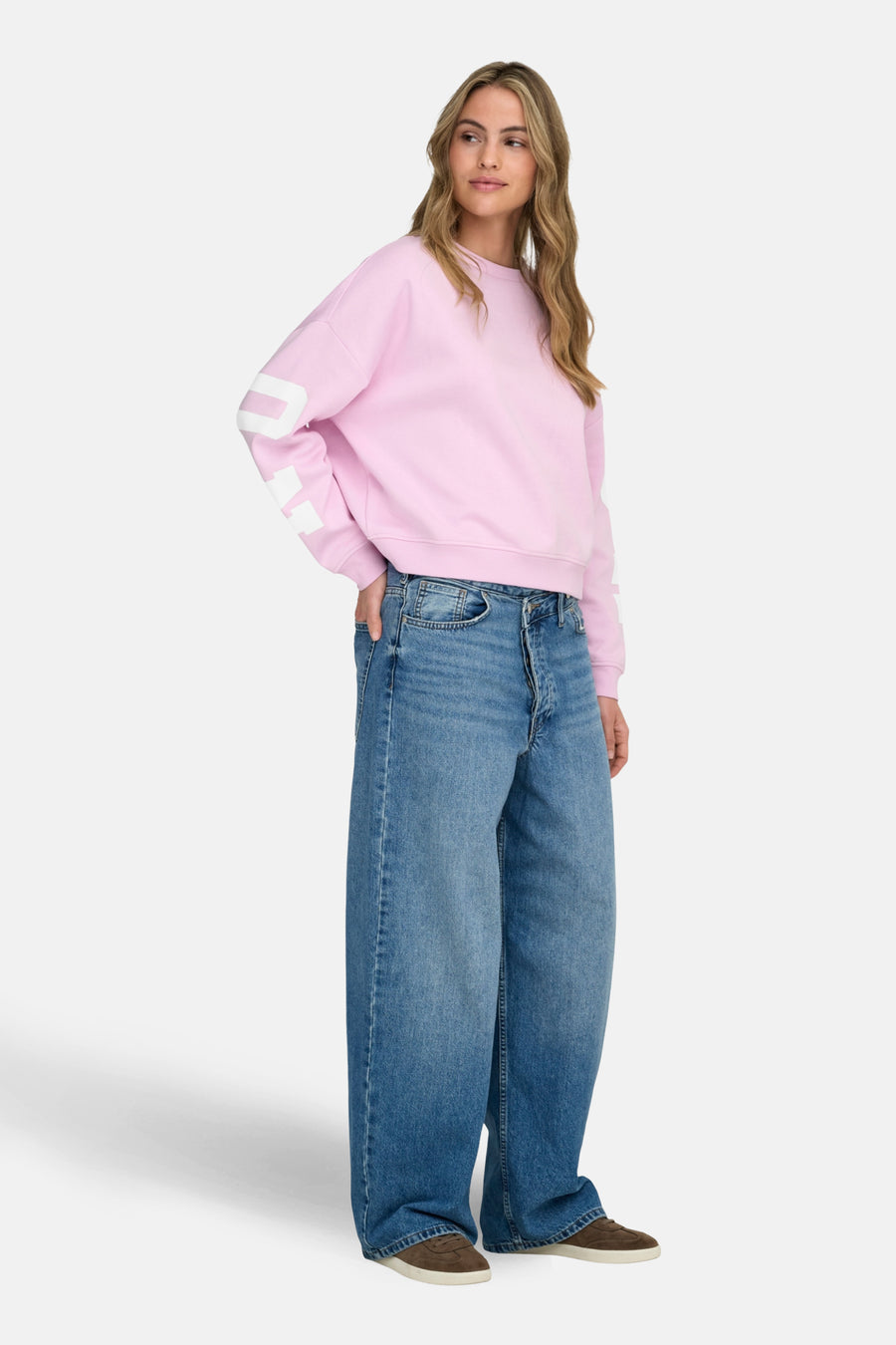 Sweat rose de ONLY, avec un lettrage blanc sur les manches, porté avec un jean large et des chaussures marron.