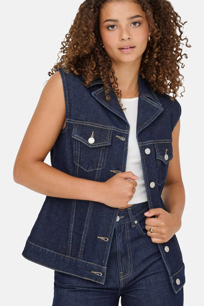 Veste sans manches en denim bleu foncé de ONLY, portée avec un haut blanc et un pantalon assorti.