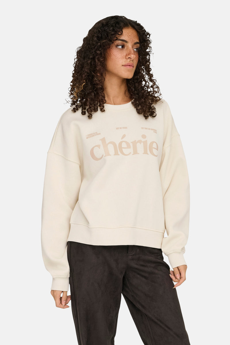 Sweat beige by ONLY®, avec inscription « chérie » sur le devant, porté avec un pantalon marron.
