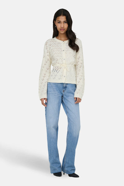 Gilet écru de ONLY, porté avec une ceinture nouée, sur un jean bleu ample et des chaussures noires.
