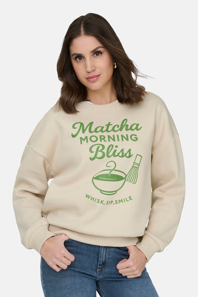 Sweat-shirt écru col rond de ONLY, avec motif imprimé thé matcha, porté avec un jean bleu.
