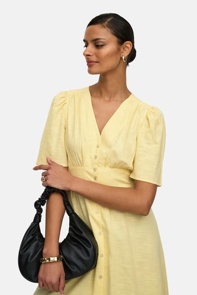 Robe jaune de JDY, avec des manches courtes bouffantes et une fermeture boutonnée, portée avec un sac à main noir et des boucles d'oreilles dorées.
