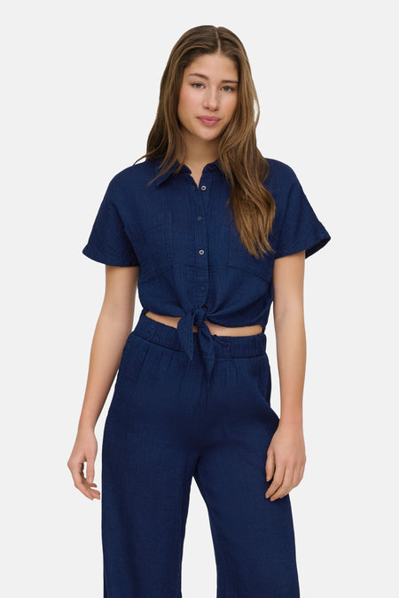 Chemisier bleu à manches courtes par ONLY®, noué à la taille, porté avec un pantalon assorti.