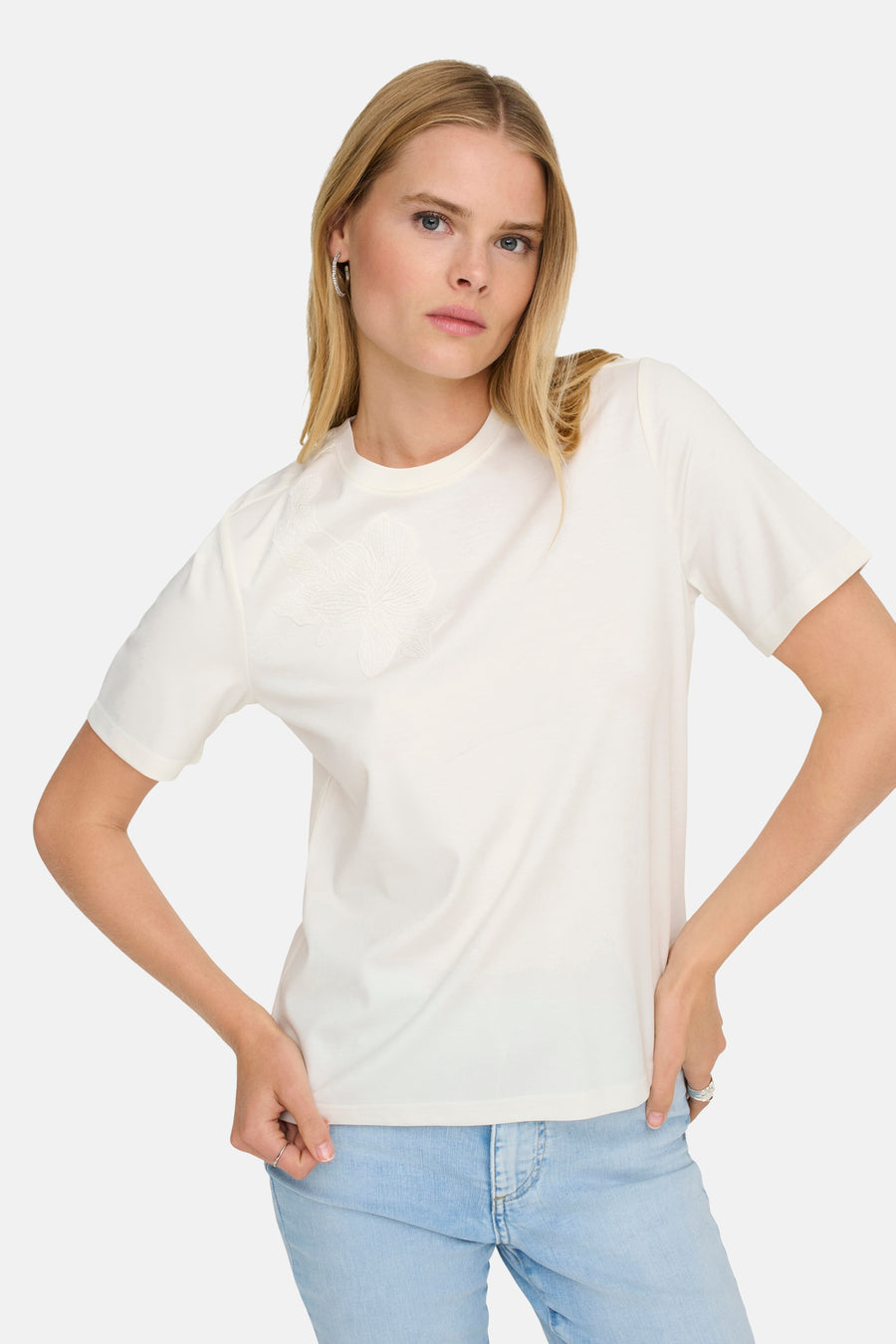T-shirt blanc à manches courtes de ONLY, avec une fleur brodée et porté avec un jean bleu clair.
