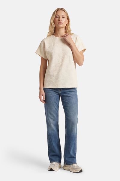 T-shirt beige à manches courtes de ONLY, avec un motif floral texturé, porté avec un jean et des baskets.
