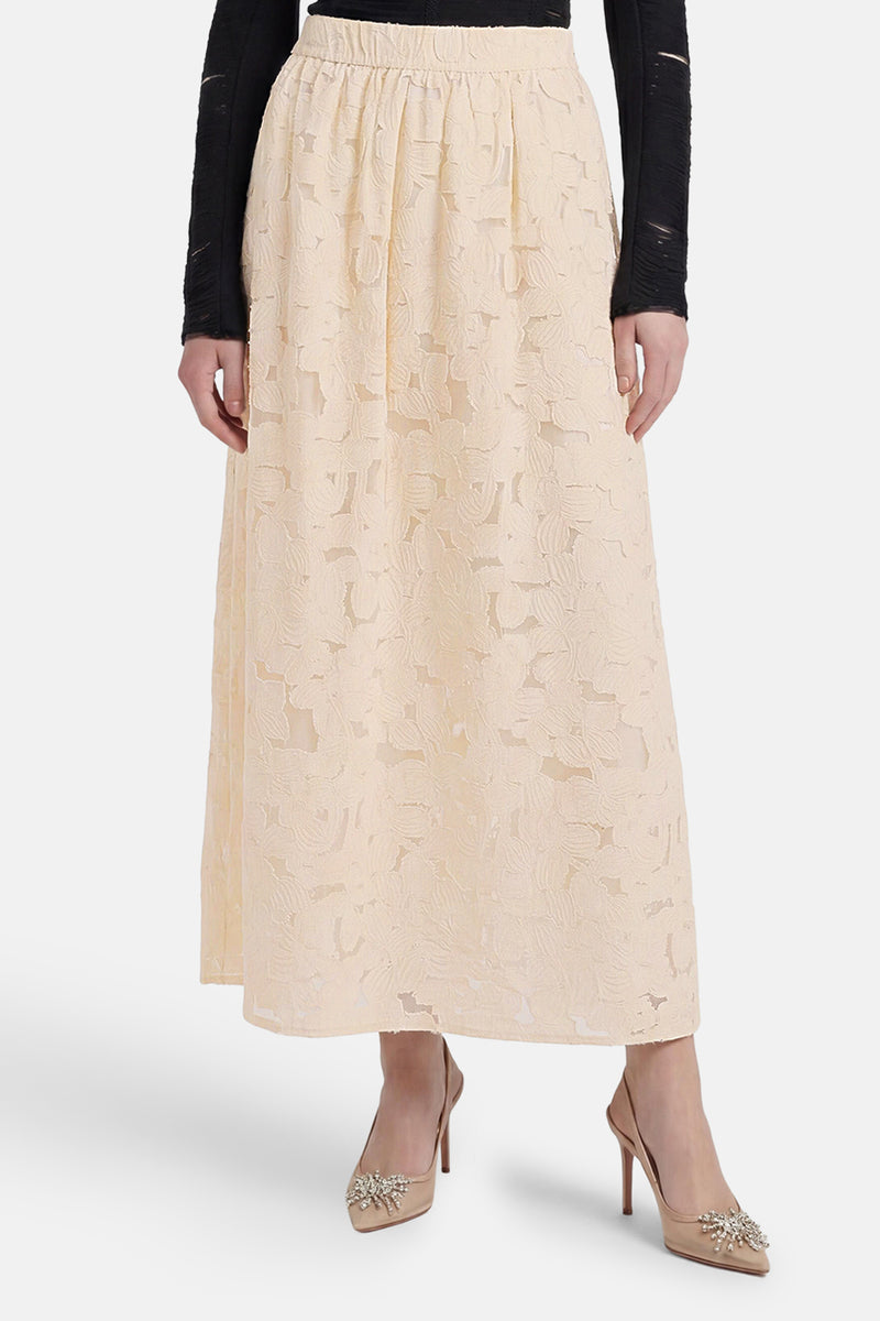 Beige long lace skirt with floral cutout pattern, elastic waist, and flowing silhouette, worn with black top and embellished heels.