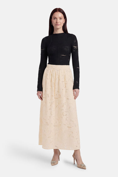 Beige long skirt with floral lace texture, elastic waist, full-length A-line silhouette, worn with black top and embellished heels, front view.