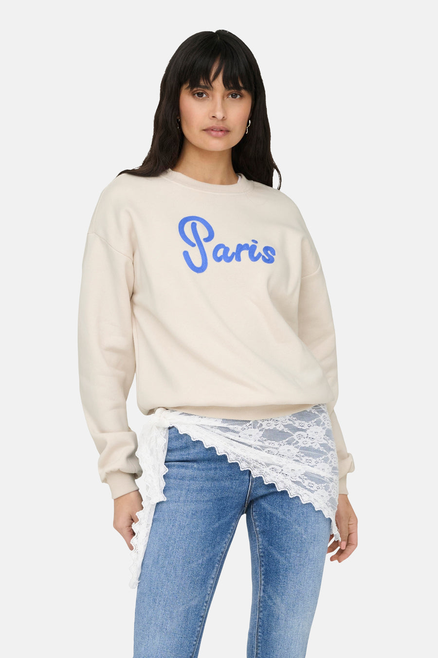 Sweat-shirt beige ONLY orné du mot « Paris » en bleu, porté avec un jean et un jupon en dentelle.
