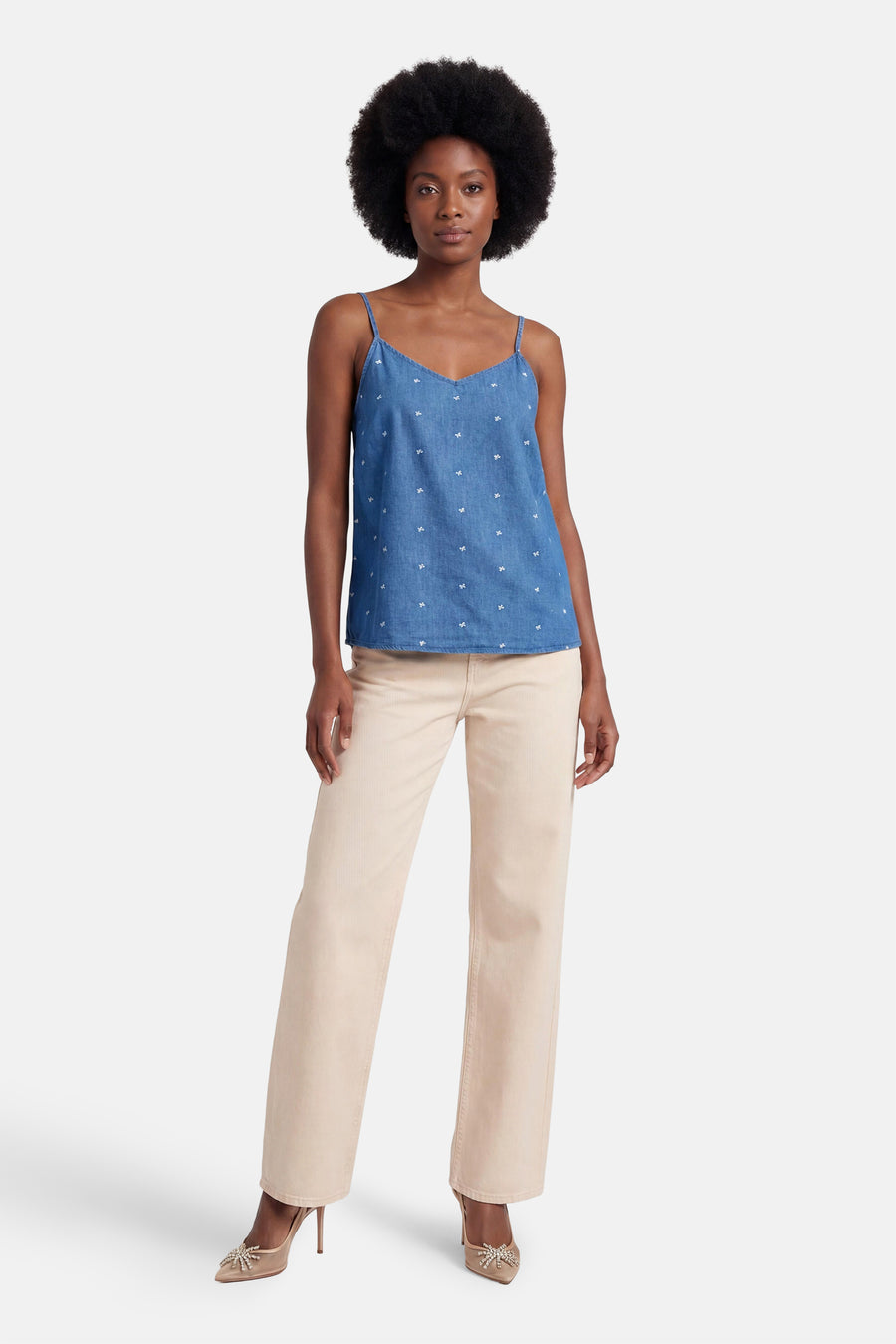 Débardeur en denim bleu moyen de ONLY® à fines bretelles, col en V et motifs de nœuds blancs, porté avec un pantalon beige.