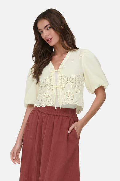 Chemise jaune pâle de ONLY® à broderie anglaise en cœurs, manches bouffantes courtes, volants et liens à nouer, portée avec un pantalon terracotta.