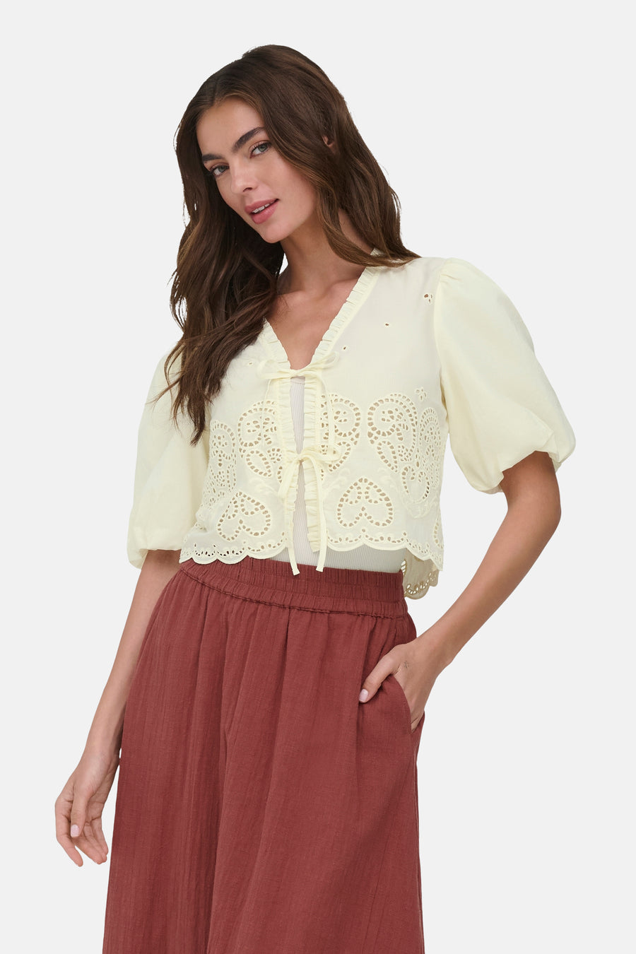 Chemise jaune pâle de ONLY® à broderie anglaise en cœurs, manches bouffantes courtes, volants et liens à nouer, portée avec un pantalon terracotta.