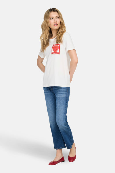 T-shirt blanc à manches courtes de ONLY, orné d'un cœur en crochet rouge, porté avec un jean bleu et des ballerines rouges.

