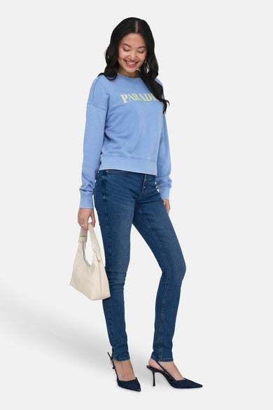Sweat bleu à col rond délavé de ONLY®, avec inscription «PARADISE» jaune, porté par une femme.