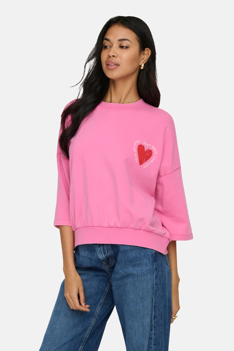 Sweatshirt van ONLY versierd met een rood hartpatroon met letters, gedragen met een spijkerbroek en beige schoenen.