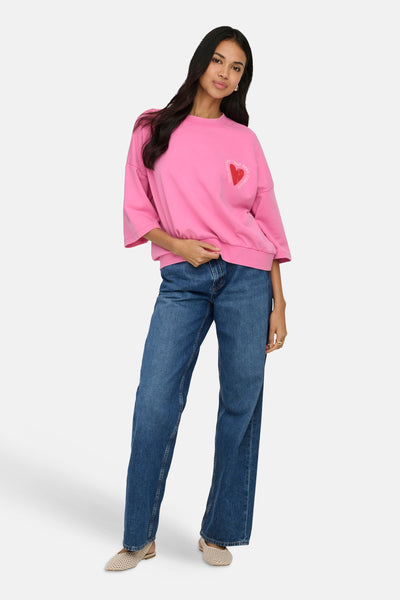 Sweat rose de ONLY orné d'un motif de cœur rouge avec des lettres, porté avec un jean et des chaussures beige.