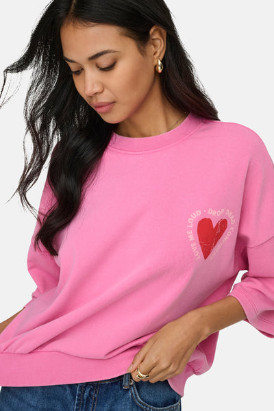 Sweat-shirt rose col rond de ONLY, avec un imprimé cœur rouge et porté sur un jean bleu.
