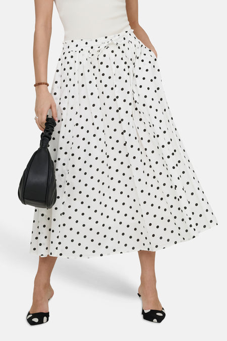 Jupe longue blanche à pois noirs de JDY, avec une ceinture nouée, portée avec un haut blanc et des mules assorties.
