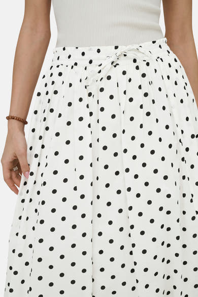 Jupe longue blanche à pois, par JDY, avec ceinture nouée et bracelet marron.
