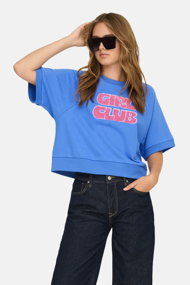 Blauwe trui van ONLY, met een roze "Girl Club" print, gedragen met een donkere flared jeans en een zonnebril.