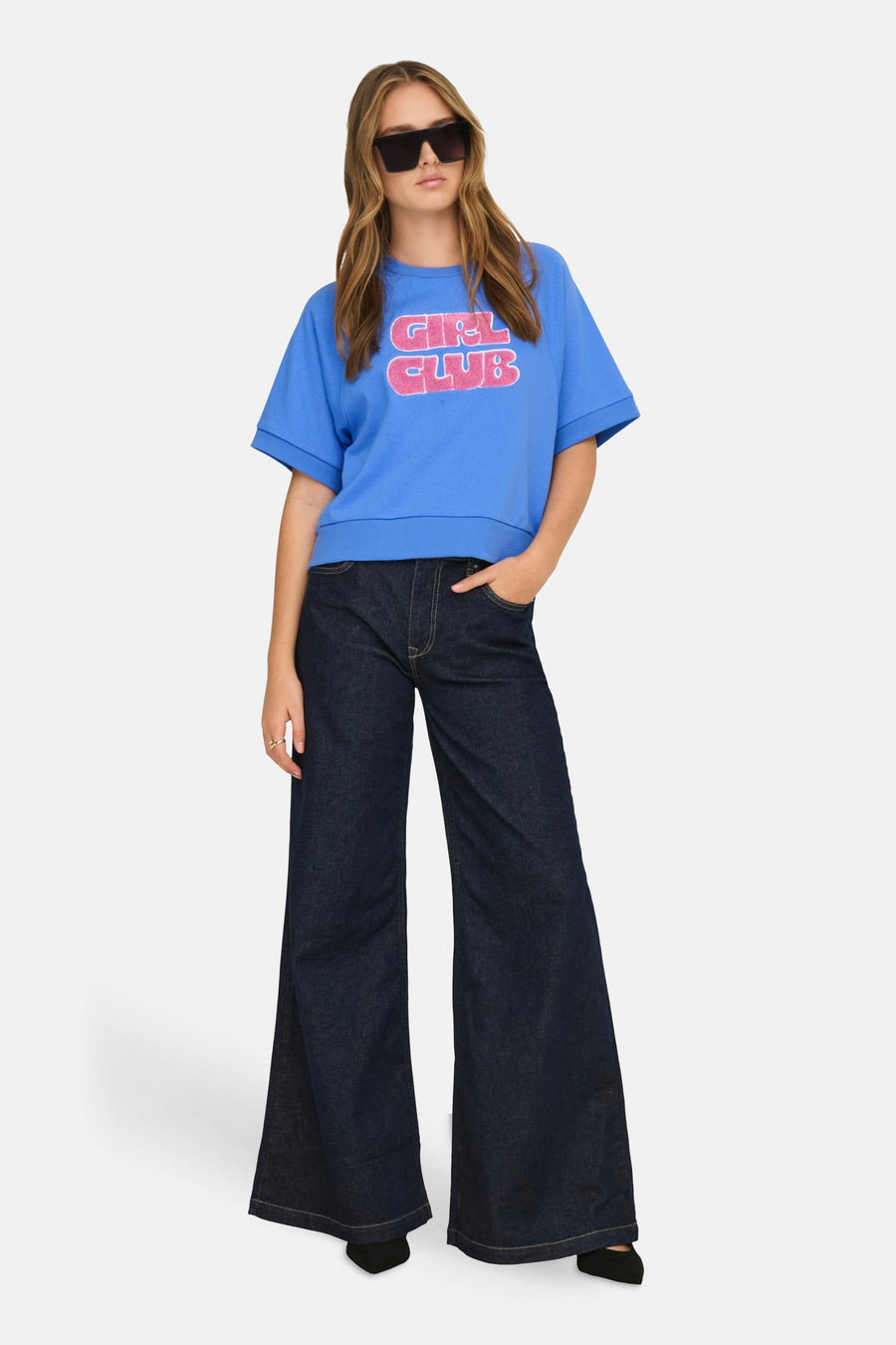 Sweat-shirt bleu de ONLY, avec un motif "Girl Club" rose, porté avec un jean évasé foncé et des lunettes de soleil.