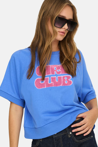Blauwe trui van ONLY, met een roze "Girl Club" print op de voorkant, gedragen met donkere jeans en vierkante zonnebril.