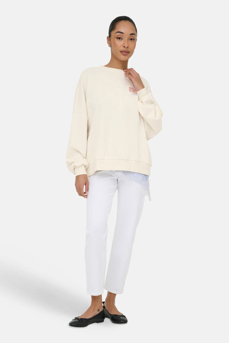 Sweat beige à col rond de ONLY® avec broderie 'Sardines' et poissons, porté avec un top en dentelle bleue, un pantalon blanc et des ballerines.