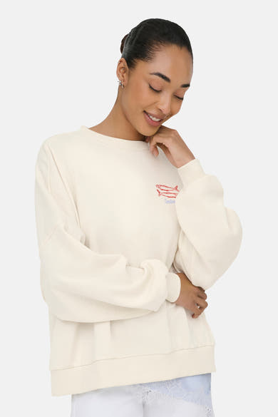 Beige sweater van ONLY® geborduurd met twee rode sardines en het woord "Sardine", gedragen door een lachende vrouw.