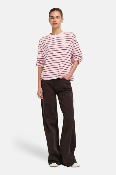 Pale lilac long-sleeve striped T-shirt with burgundy horizontal stripes, worn loose with rolled sleeves and paired with dark wide-leg pants and white shoes.