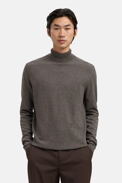 Pull à col roulé brun par SELECTED, porté avec un pantalon marron uni, les mains dans les poches.

