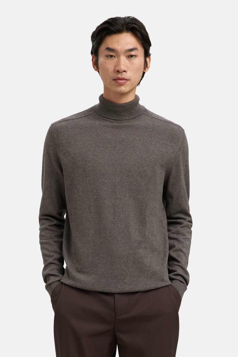 Pull à col roulé brun par SELECTED, porté avec un pantalon marron uni, les mains dans les poches.
